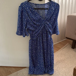 Blå blommig kortklänning med v-ringning - Supersöt blå kortklänning med vitt blommönster och v-ringning. Klänningen har korta, vida ärmar och en twistad detalj framtill som ger en snygg siluett. Perfekt för varma dagar och har en luftig känsla tack vare det tunna materialet.