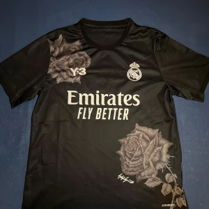 Real Madrid x Y-3 svart fotbollströja - Svart Real Madrid x Y-3 fotbollströja från Adidas med stora gråa rosor tryckta på axel och nederkant. Tröjan har korta ärmar, rund hals och är tillverkad i lätt, ventilerande polyester. Framsidan har klubbmärke, Y-3-logga och Emirates Fly Better-tryck.
