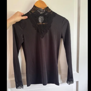 Svart ribbad blus med spets från Jacqueline de Yong - Supersnygg svart ribbad blus från Jacqueline de Yong i storlek S. Hög hals med detaljerad spets framtill och vid ärmsluten. Långärmad och figurnära passform, perfekt för dig som gillar stilrena men ändå unika plagg.
