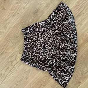 Leopardmönstrad kortkjol från D-XEL - Snygg kortkjol från D-XEL med leopardmönster i beige, brunt och svart. Kjolen har volangdetaljer och ett glansigt, lätt material som ger en cool och trendig look. Perfekt för dig som vill sticka ut med en djurisk vibe. Storlek 140, 10åring.