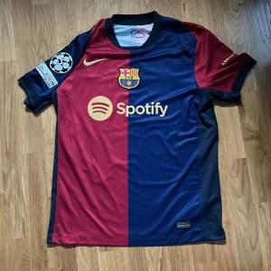 FC Barcelona Lamine Yamal matchtröja - Snygg FC Barcelona fotbollströja med Lamine Yamal och nummer 19 på ryggen. Tröjan är kortärmad och har klassisk halva-röd halva-blå design, klubbmärke, Champions League-märke. Tillverkad i lätt och ventilerande polyester. Det är en Replika! Handtvätt rekomenderas.