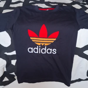 Svart Adidas t-shirt med röd logga - Svart t-shirt från Adidas med stor broderad trefoil-logga i rött och orange på bröstet samt vit Adidas-text. Klassisk rund halsringning med röd detalj på insidan. Skön passform och tillverkad i mjuk bomull.