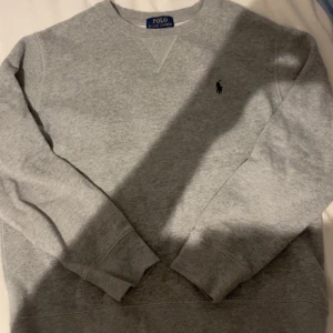 Grå sweatshirt från Polo Ralph Lauren - Klassisk grå sweatshirt från Polo Ralph Lauren med rund hals och broderad marinblå logga på bröstet. Tröjan har långa ärmar och ribbade muddar vid ärmslut och nederkant. Perfekt för en chill och stilren look.