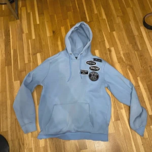 Ljusblå hoodie från Mercier - Säljer en ljusblå hoodie från Mercier med huva och snörning. Tröjan har flera svarta och vita patchar på bröstet och en stor magficka. Perfekt för dig som gillar streetwear och vill ha en chill look.