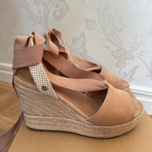 UGG sandaler med kilklack och ballerinaknytning - Snygga sandaler från UGG i ljusrosa/beige mocka med breda band att knyta runt ankeln. Skorna har hög kilklack i flätad jute och öppen tå. Perfekta för sommaren med sin ljusa färg och trendiga espadrille-vibe. Jättefint skick, använd ett par gånger. Modell: W Shiloh, storlek 39.
