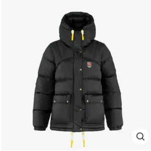 Svart dunjacka från Fjällräven - Svart dunjacka från Fjällräven med huva och gula detaljer på snörningen. Jackan har två stora fickor framtill, tryckknappar och dragkedja. Klassisk Fjällräven-logga på bröstet. Perfekt för kalla dagar när du vill hålla dig varm och snygg.