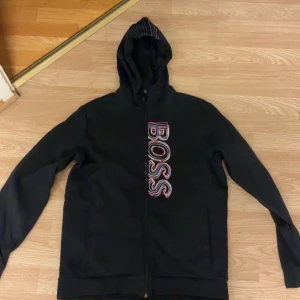 Svart hoodie från BOSS med dragkedja - Svart hoodie från BOSS med stor, färgglad BOSS-logga vertikalt på framsidan. Tröjan har huva, långa ärmar och hel dragkedja. Perfekt för dig som gillar streetstyle och vill ha en snygg och bekväm look.