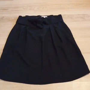 Svart kortkjol med knyt från H&M - Svart kortkjol från H&M med bred midja och knytband. Kjolen har dragkedja i sidan och lätt utställd form. Perfekt för dig som gillar en enkel men snygg stil.