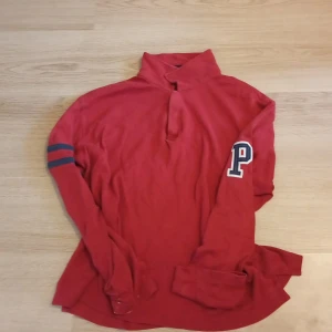 Röd Peak Performance pikétröja L - Röd långärmad pikétröja från Peak Performance med hög krage och broderad P på ärmen. Två mörkblå ränder på ena ärmen ger en sportig vibe. Skön bomullskvalitet och relaxed fit, perfekt till jeans eller chinos.