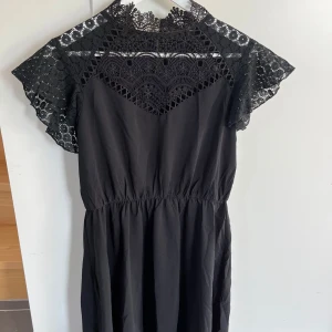 Svart spetsklänning från SHEIN - Supersnygg svart kortklänning från SHEIN med vacker spets upptill och på ärmarna. Klänningen har elastisk midja, korta ärmar och hög hals med spetsdetaljer. Perfekt för dig som vill ha en feminin och trendig look.