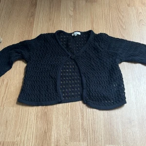 Svart stickad kofta från SAMSA Young - Svart stickad kofta från SAMSA Young med lång ärm och ett snyggt virkat mönster. Koftan har v-ringning och knäpps med en knapp framtill. Perfekt att slänga över en t-shirt för en chill look.