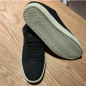 Svarta sneakers från Saint Laurent - Snygga svarta sneakers från Saint Laurent med vit sula och klassisk snörning. Skorna har en stilren design med rund tå och diskret logga på sidan. Ovandelen är i textil och sulan i gummi, perfekt för en clean och modern look.