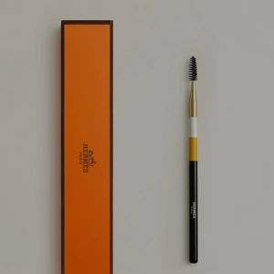 Hermès ögonbrynsborste med ask - Lyxig ögonbrynsborste från Hermès med svart, vitt och gult handtag samt guldfärgad topp. Kommer i ikonisk orange Hermès-ask. Perfekt för att forma och styla brynen med stil.
