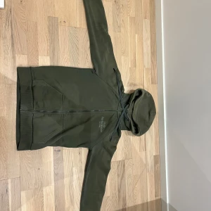 Olivgrön hoodie Peak Performance S - Olivgrön hoodie från Peak Performance med broderad logga på bröstet. Tröjan har dragkedja, huva med snörning och två fickor framtill. Insidan är mjuk och fleecefodrad, perfekt för kyliga dagar. Klassisk och stilren design som funkar året runt.