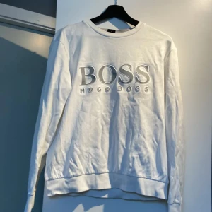 Vit slim fit tröja från Hugo Boss - Vit långärmad tröja från Hugo Boss med stort BOSS-tryck framtill. Slim fit-modell med ribbade muddar vid ärmslut och nederkant. Tillverkad i mjuk bomull, perfekt för en clean och stilren look.