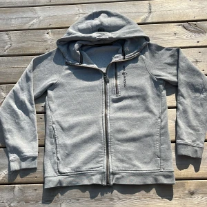 Grå hoodie från Sail Racing med dragkedja - Snygg ljusgrå hoodie från Sail Racing med hel dragkedja framtill och extra bröstficka med dragkedja. Tröjan har huva, två sidofickor och är tillverkad i mjuk bomullsblandning. Perfekt för kyliga dagar och enkel att matcha med jeans eller joggers.