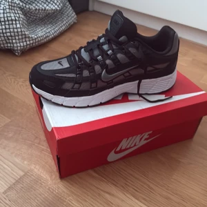 Nike P-6000 svart/grå sneakers - Nike P-6000 sneakers i svart och grått med vit sula. Skorna har meshpaneler och detaljer i syntet, klassisk snörning och en sportig vibe. Perfekta för dig som gillar streetstyle och vill ha en bekväm och snygg sneaker till vardags.