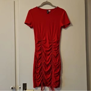Röd kortklänning från H&M Divided - Snygg röd kortklänning från H&M Divided med korta ärmar och tight passform. Klänningen har coola rynkade detaljer och snörning framtill som ger en trendig look. Tillverkad i stretchigt material av polyester och elastan. Perfekt för dig som gillar att sticka ut.