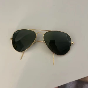 Ray-Ban Aviator guld solglasögon - Snygga Ray-Ban Aviator solglasögon med tunna guldfärgade metallbågar och mörka linser. Klassisk pilotform som alltid är trendig. Modellnummer RB 6049, storlek 55 mm. Perfekta för att lyfta din stil i sommar.