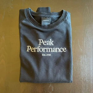 Peak Performance - Svart sweatshirt från Peak Performance med vit broderad logga och text på bröstet. Klassisk rund halsringning och långa ärmar med ribbade muddar. Perfekt för dig som gillar en clean och sportig stil. 