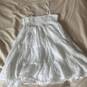 Vit somrig klänning från Gina Tricot - Superfin vit klänning från Gina Tricot med smock upptill och tunna axelband. Klänningen har en luftig, volangig kjol i flera lager och är perfekt för varma dagar. Tillverkad i ett lätt och svalt material som känns skönt mot huden.