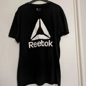 Svart Reebok t-shirt Supremium tee - Svart t-shirt från Reebok med stor vit logga och text på bröstet. Klassisk passform med rund hals och korta ärmar. Härlig träningstshirt i modellen Supremium tee. Kan förekomma lite ytnoppor, men den är i väldigt bra skick. 
