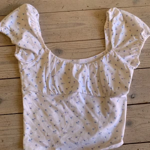 Vit blommig blus från Hollister - Denna topp har använts ett fåtal gånger. Supersöt vit blus från Hollister med små blå blommor och puffiga korta ärmar. Toppen har en söt rynkad detalj vid bysten och en feminin, lätt croppad passform.