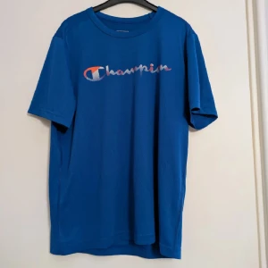 Blå Champion t-shirt  - Snygg blå träningst-shirt från Champion med klassisk logga i vitt, rött och blått framtill. Tillverkad i lätt och luftig polyester, perfekt för träning eller chill. Normal passform och korta ärmar. Enkel och sportig stil med rund halsringning.