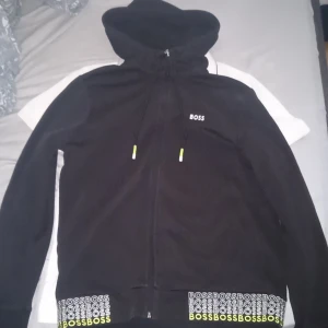 Svart hoodiejacka från BOSS - Svart hoodiejacka från BOSS med dragkedja, snörning i huvan och tryckt BOSS-logga i vitt och gult längs nederkanten. Jackan har långa ärmar och en clean look med små detaljer i neon. Perfekt för en chill och sportig stil.
