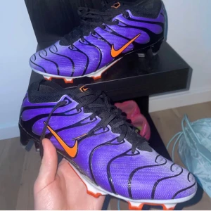 Nike tn elite - Snygga fotbollsskor från Nike i lila med svarta och orange detaljer. Skorna har en cool vågig design, svart snörning och orange swoosh på sidan. Yttersulan är vit med orange dobbar, perfekt för dig som vill sticka ut på planen.