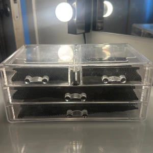 Transparent sminkförvaring med lådor - Snygg och praktisk sminkförvaring i genomskinlig akryl med fyra lådor, perfekt för att organisera sminkborstar, läppstift eller ögonskuggor. Svarta mattor i varje låda håller sminket på plats och ger en stilren look till sminkbordet.