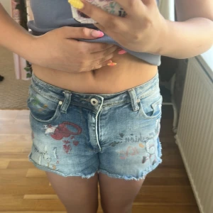 Jeansshorts med tryck från Miss Two - Snygga blå jeansshorts från Miss Two med coola tryck och slitna detaljer. Shortsen har råa kanter, klassisk femficksmodell och midwaist passform. 