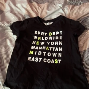 Svart t-shirt med tryck från H&M - Svart t-shirt från H&M med coolt tryck i vitt och neongult framtill. Texten har New York-vibe och t-shirten har klassisk rund hals och korta ärmar. Perfekt för dig som gillar streetstyle och enkel design. Materialet är mjuk bomull som är skön mot huden.