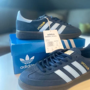 Adidas Handball Spezial marinblå sneakers - Adidas Handball Spezial sneakers i marinblå mocka med klassiska vita ränder och guldfärgad Spezial-logga på sidan. Snygg retrostil med svart sula och snörning. Perfekta för dig som gillar streetwear och tidlös design.