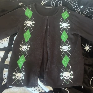 Svart kofta med döskallemönster Liquorbrand - Svart kofta från Liquorbrand med gröna och vita argyle-rutor och döskallemönster längs fram och på ärmarna. Rund halsringning, knäppning framtill och långärmad. Perfekt för dig som gillar alternativ stil och vill sticka ut.