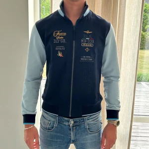 Aeronautica Militare jacka blå - Snygg bomberjacka från Aeronautica Militare med mörkblå kropp och ljusblå ärmar. Jackan har broderade detaljer och patchar framtill, dragkedja och ribbade muddar. Perfekt för dig som gillar unik stil och italiensk flyginspiration.