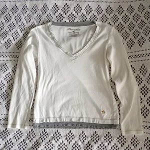 Vit tröja Abercrombie - Vit vintage supersöt långärmad tröja från Abercrombie i 100% bomull. V-ringad med grå kant och råa kanter vid halsen. Liten guldfärgad älg broderad nertill. Perfekt basic med snygga detaljer och skön. Står L men skulle säga S (säljer även en likadan grå!)🩷