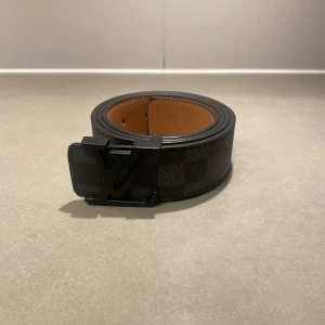 Svart LV bälte - Snyggt Louis Vuitton bälte i svart och grått Damier Graphite-mönster med klassiskt LV-spänne i svart metall. Bältet är gjort i exklusivt skinn och har en stilren, fyrkantig form. Insidan är ljusbrun och mjuk. Perfekt accessoar för att lyfta din outfit.