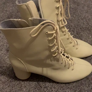 Beige snörade boots från Zara - Snygga beige boots från Zara Trafaluc med snörning hela vägen upp och rund tå. Skorna har en stabil klack och är gjorda i syntetmaterial. Perfekta för dig som vill ha en stilren och trendig look.