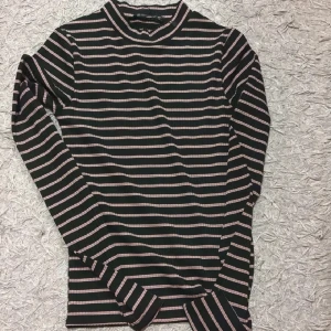 Randig svart och rosa långärmad tröja - Snygg långärmad tröja med svarta och ljusrosa horisontella ränder. Tröjan har en högre rundad hals och är ribbad i materialet, vilket ger en skön passform. Perfekt för dig som gillar en enkel men trendig stil.