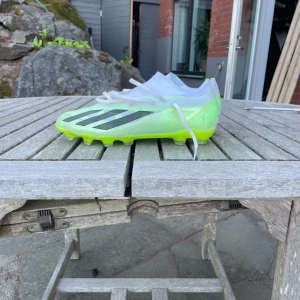 Adidas fotbollsskor i neon och vitt - Snygga Adidas fotbollsskor i vitt och neon-gult med svarta ränder på sidan. Skorna har snörning, platt sula med dobbar och är tillverkade i syntetmaterial. Perfekta för dig som vill sticka ut på planen med en modern och sportig look. Väldigt bra skick endast använd en match och sedan gjorde de för ont så fick köpa nya😊