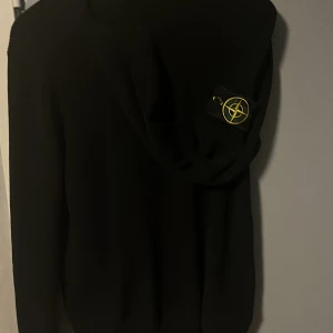 Svart ribbad tröja från Stone Island - Säljer en stilren svart ribbad tröja från Stone Island med deras ikoniska märke på ärmen. Tröjan har långa ärmar och en klassisk passform. Perfekt för en cool och avslappnad stil.