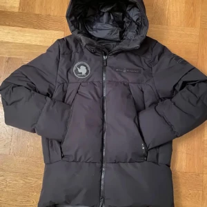 Svart dunjacka från Sail Racing - Säljer en svart dunjacka från Sail Racing med huva och dragkedja. Jackan har en stilren design med en logotyp på bröstet och armen. Perfekt för kyliga dagar.