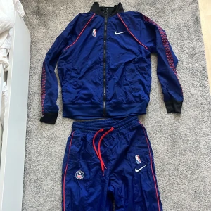 Blå tracksuit från Nike med NBA-logga - Snygga blå tracksuit från Nike med röd snörning och NBA-logga. Byxorna har elastiska muddar och en sportig look med röda detaljer längs sidorna. Perfekta för basket eller annan träning.