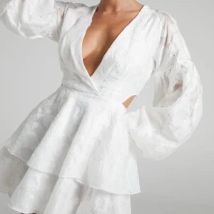 Vit studentklänning med volanger - Elegant vit klänning från showpo med v-ringad hals och volanger. Helt ny och prislappen är på. Klänningen har långa ärmar och ett omlottmönster som ger en stilfull look. 