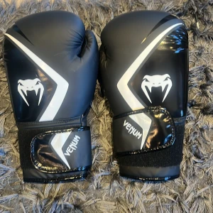 Svarta boxningshandskar från Venum - Snygga svarta boxningshandskar från Venum med vit logga och detaljer. De har en justerbar kardborrestängning för bra passform och stöd. Perfekta för träning och sparring. 🥊