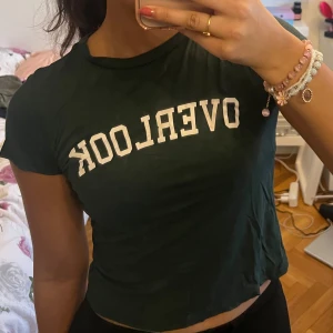Mörkgrön t-shirt med text - Säljer en mörkgrön t-shirt med texten 'OVERLOOK' i vitt på framsidan. T-shirten har en normal passform och korta ärmar. Perfekt för en avslappnad stil.