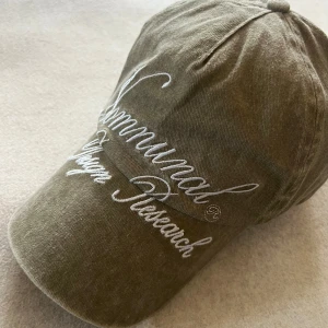 Faded Khaki Embroidered Logo Cap - Denna justerbara khakifärgade keps har ett broderat logomärke både på ramen och framsidan, vilket ger den en stilren och unik look. Oanvänd och i perfekt skick, erbjuder den en bekväm passform och en diskret men distinkt detalj till din outfit. Idealisk för både vardag och sportiga tillfällen