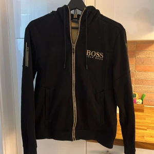 Svart hoodie/dragkedja från Hugo Boss (Strl S) - Snygg svart jacka från Hugo Boss med dragkedja och logotyp på bröstet. Jackan har långa ärmar och stilrena detaljer i guld. Perfekt för en trendig look. Nypris 2000kr. Säljer nu för 500kr fint skick! Det är en ganska tajt strl S, skulle säga att man kan bära den i Xs med. 