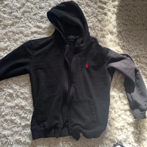 Svart hoodie från Ralph Lauren - Säljer en stilren svart hoodie från Ralph Lauren med dragkedja och huva. Den har en röd broderad logotyp på bröstet och praktiska fickor framtill. Perfekt för en avslappnad look.